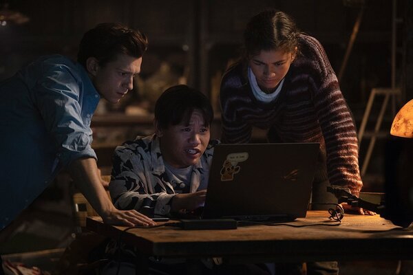 Peter (Tom Holland), Ned (Jacob Batalon), and MJ (Zendaya) are bent over a desk peering at a laptop screen. The laptop is facing away from the camera.