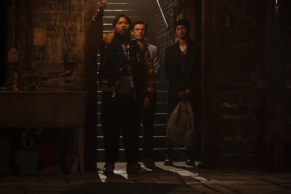 Ned (Jacob Batalon), Peter (Tom Holland), and MJ (Zendaya) are standing at the bottom of a staircase, in room with brick walls.